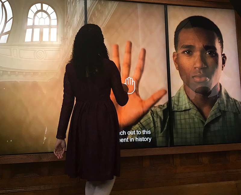 A young woman with her back to the camera touches a photo on a large touch screen of a young man with his hand raised 