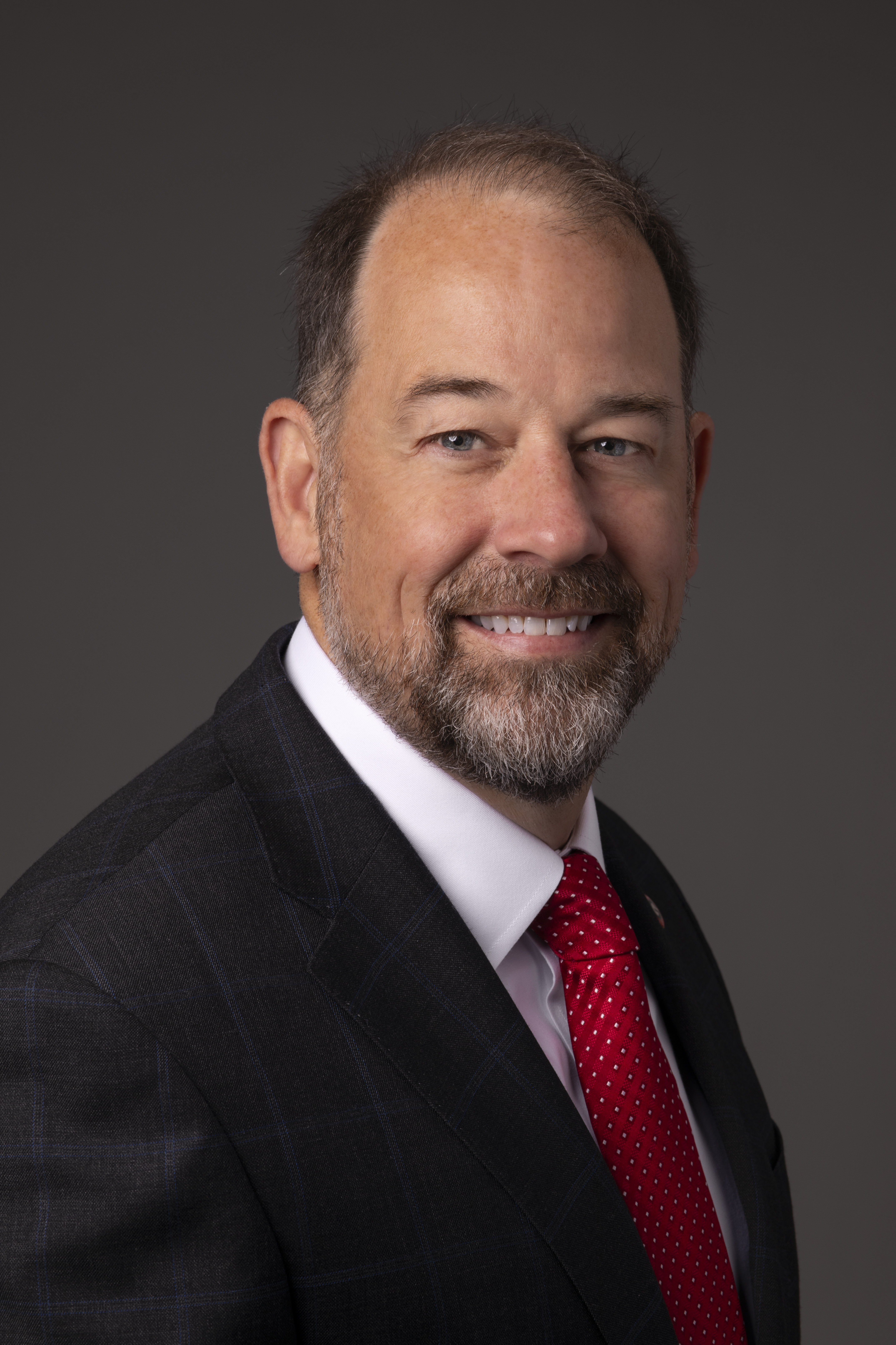 Man with short dark hair and a beard wearing a dark suit, white shirt, and red tie.