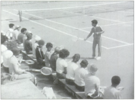A group of players sit and watch as a tennis instructor demonstrates a shot