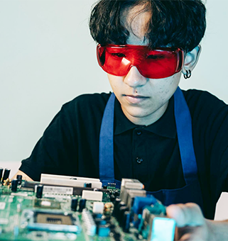 student wearing plastic red protection glasses and blue apron holding out a circuit board project